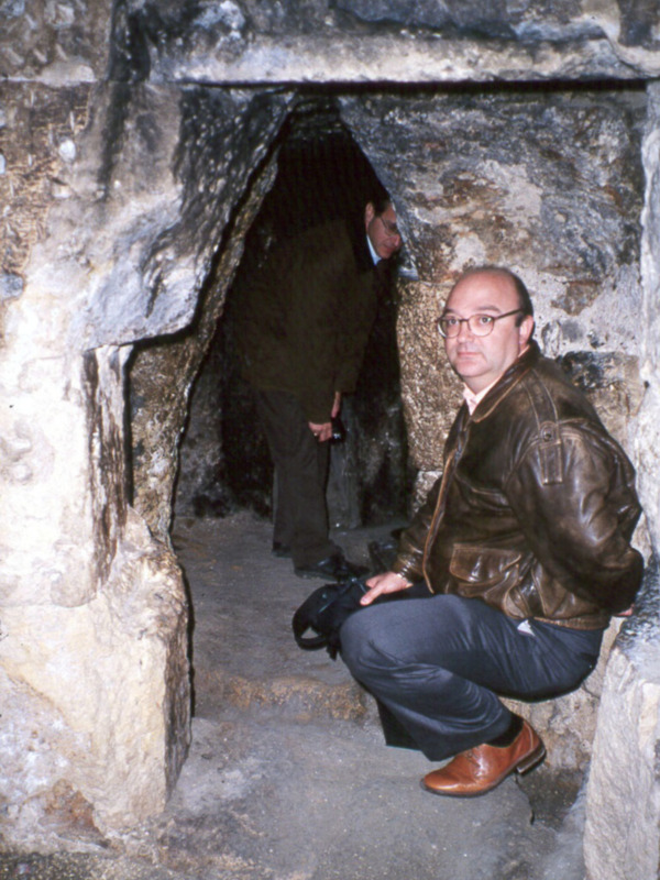Hombre sentado en una cueva antigua, con otra persona en el fondo.