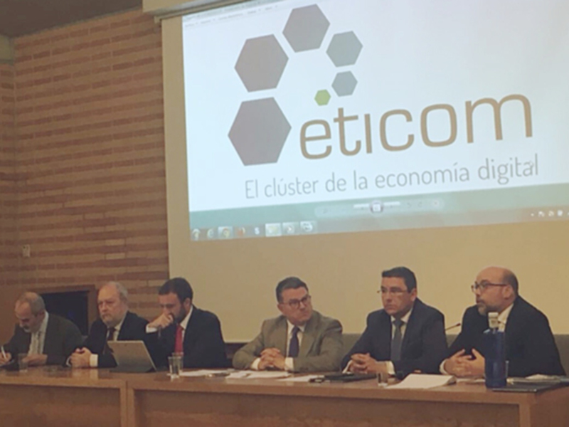 Presentación en conferencia sobre el clúster de la economía digital, con un panel de expertos sentados en una mesa con documentos y un proyector en el fondo mostrando el logo de la organización.