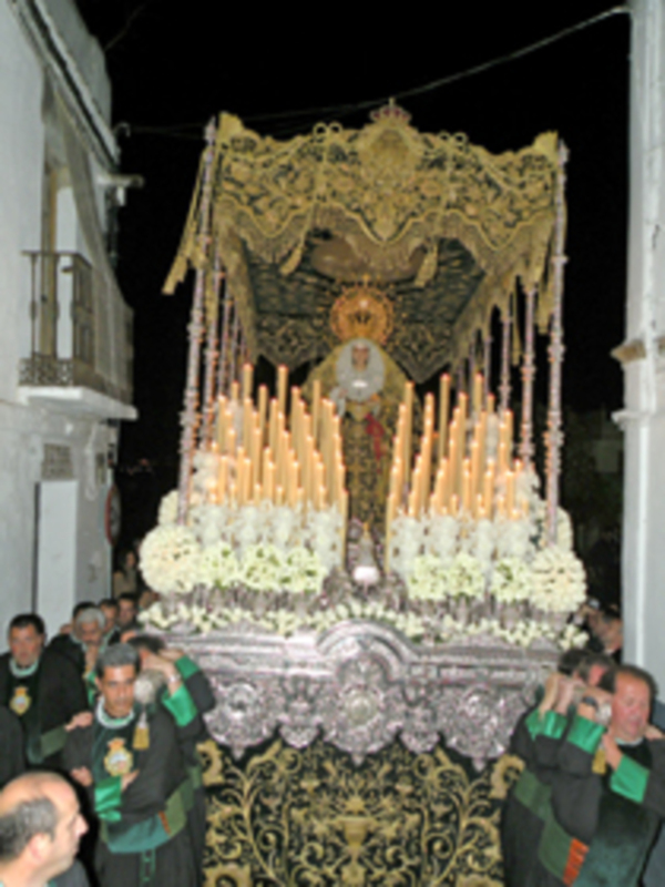 Procesión nocturna con una imagen procesional ornada con velas y flores, llevada por hombres en trajes tradicionales.