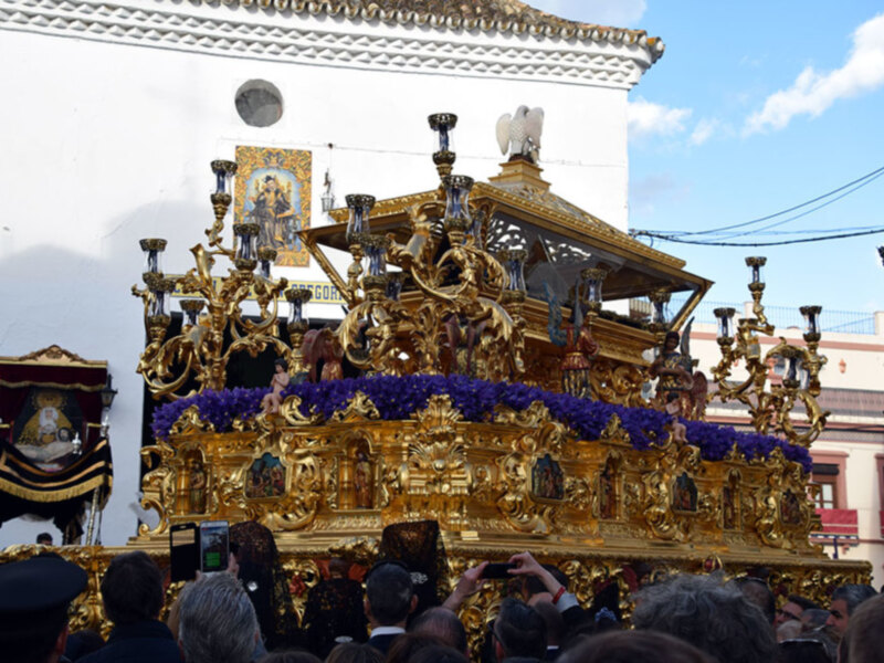 Procesión con carroza dorada y flores púrpuras en una calle de España.