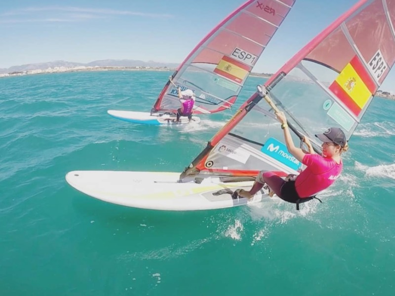 Dos personas windsurfando en aguas turquesas con banderas españolas en sus velas.