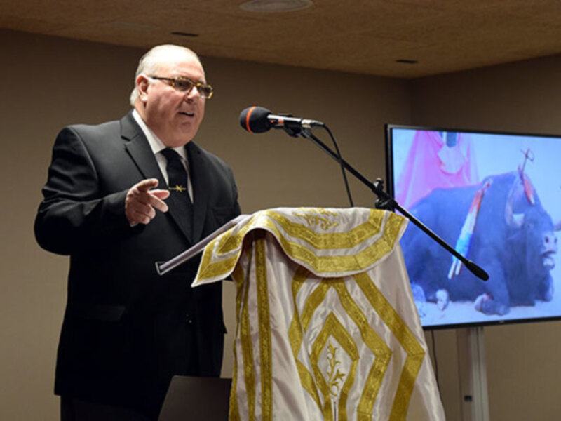 Un hombre en traje hablando en un podio con un micrófono, con una imagen de un toro en una pantalla de fondo.