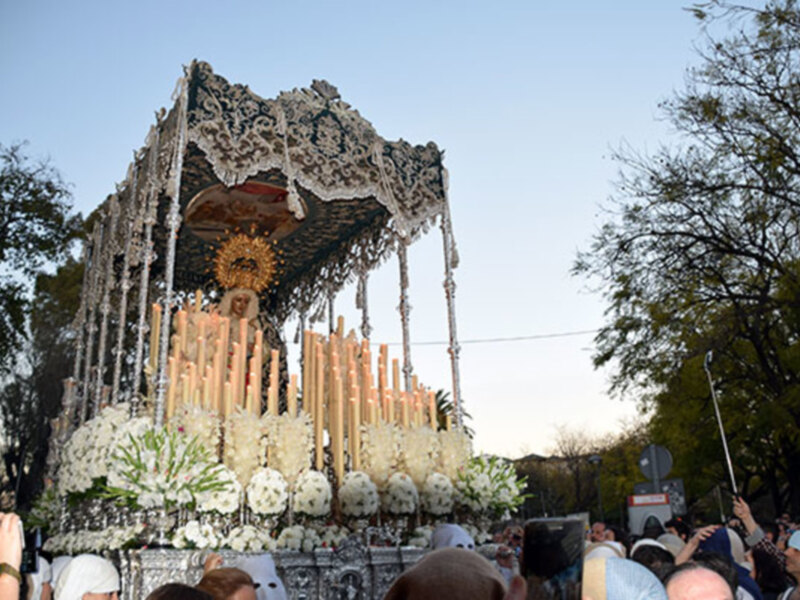Procesión religiosa con imagen de la Virgen en un paso ornado con flores y candelabros, rodeada por espectadores en una calle urbana.