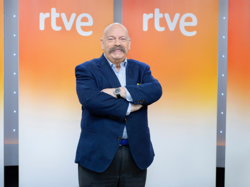 Un hombre con barba y bigote, vistiendo una chaqueta azul y una camisa azul con cuadros, posa con los brazos cruzados en frente de una pared con el logo de RTVE.