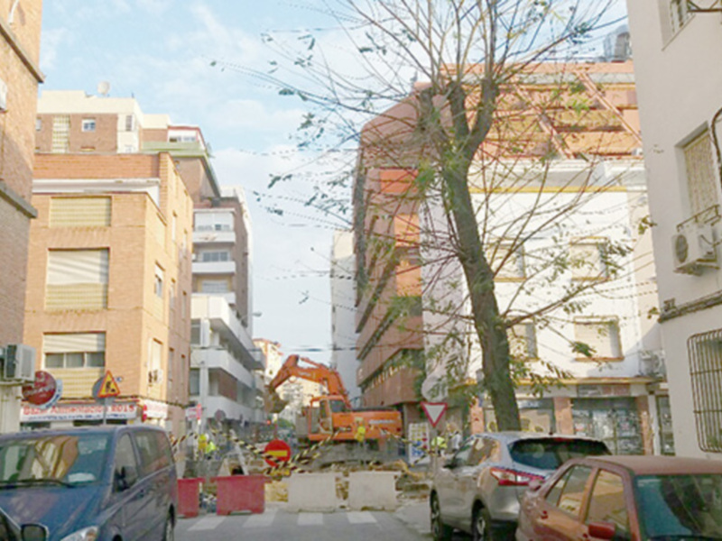 Vista de una calle urbana con edificios residenciales, un árbol y un vehículo excavador en obras de construcción en el fondo.