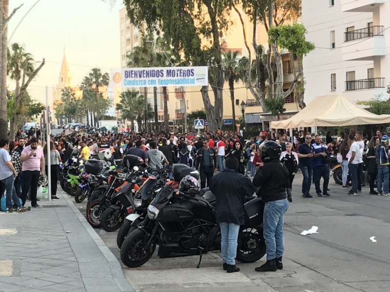 Motos estacionadas en la calle con personas de pie alrededor, y un cartel que dice 'BIENVENIDOS MOTEROS' en el fondo.