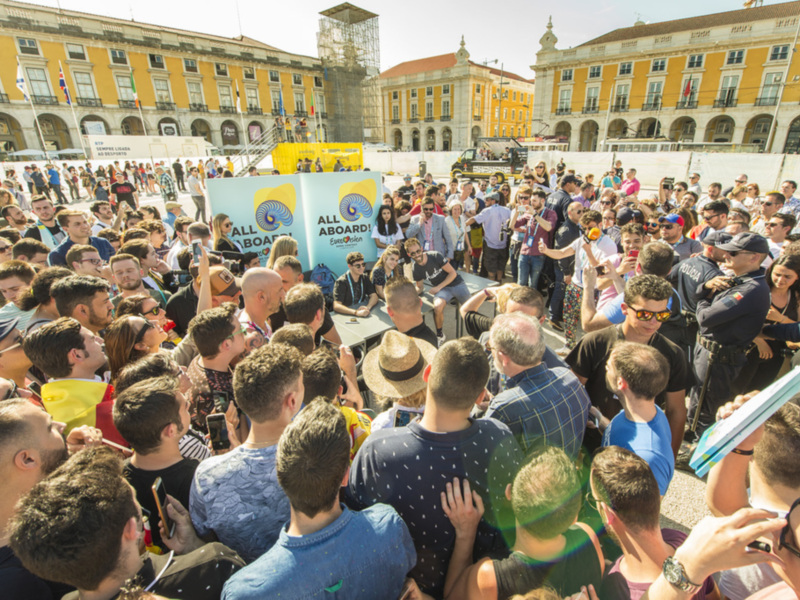 Una multitud de personas en un evento al aire libre, con una bandera azul y amarilla que dice "All Aboard" en el centro. La imagen muestra una mezcla de personas con diferentes estilos, algunas con sombrillas y otros con cascos. La escena está en un entorno urbano, con edificios históricos al fondo.