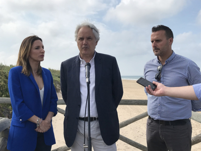 Tres personas en una playa hablando con un micrófono y un teléfono móvil, con el mar y el cielo de fondo.