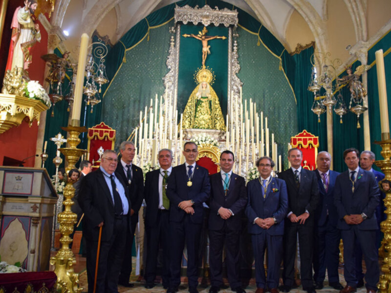 Grupo de hombres en trajes formales posando frente a una imagen religiosa en un ambiente ceremonial.