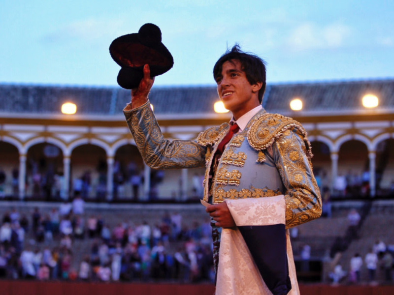 Un torero en el centro de la plaza de toros, con su capote en alto y una sonrisa en su rostro, mientras la gente en el fondo observa la celebración.