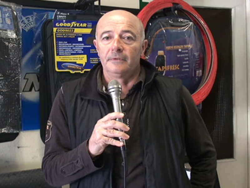 Hombre hablando con un micrófono en un entorno de taller con productos de neumáticos en el fondo.