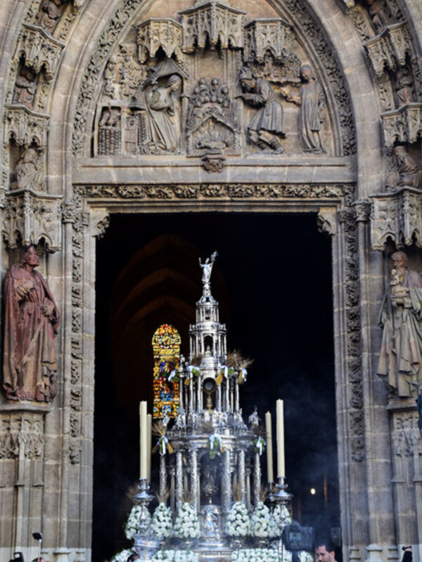 Procesión religiosa con estandarte ornado y arco gótico decorado con esculturas en una catedral.
