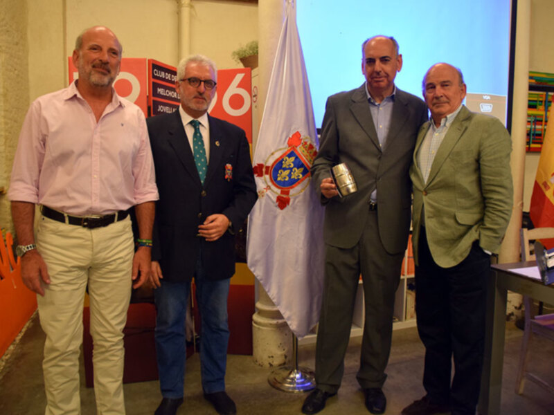 Cuatro hombres posando juntos en un evento formal con banderas y carteles de fondo.