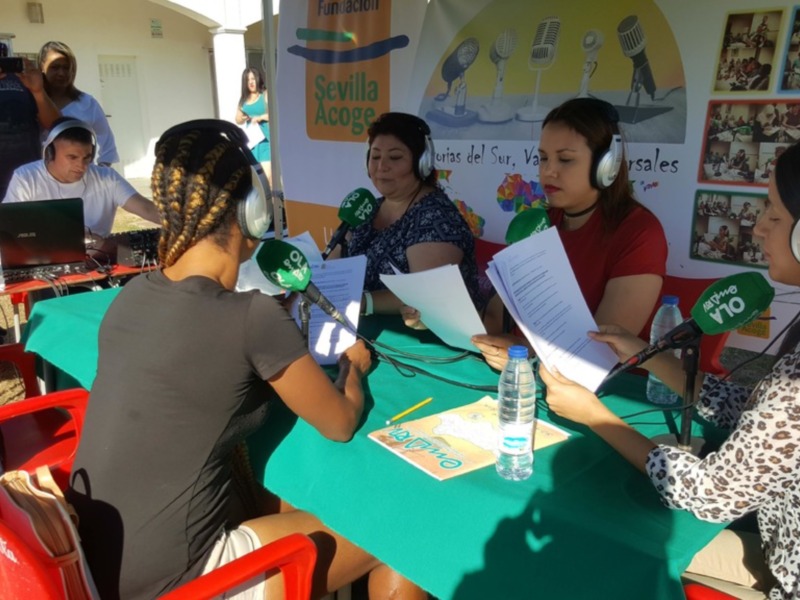 Participantes en una emisión de radio al aire libre, con micrófonos y documentos, promoviendo la Fundación Sevilla Acoge.