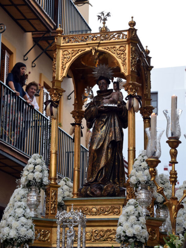 Procesión religiosa con estandarte dorado y estatua de la Virgen con el Niño, adornado con flores y candelabros.