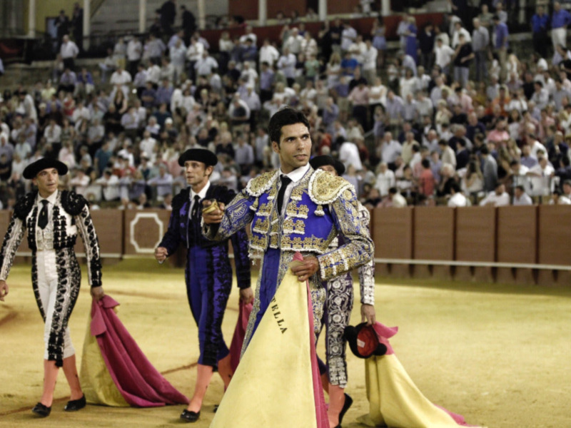 Tres matadores de toros en trajes de lujo caminan por la plaza de toros ante un público entusiasmado.