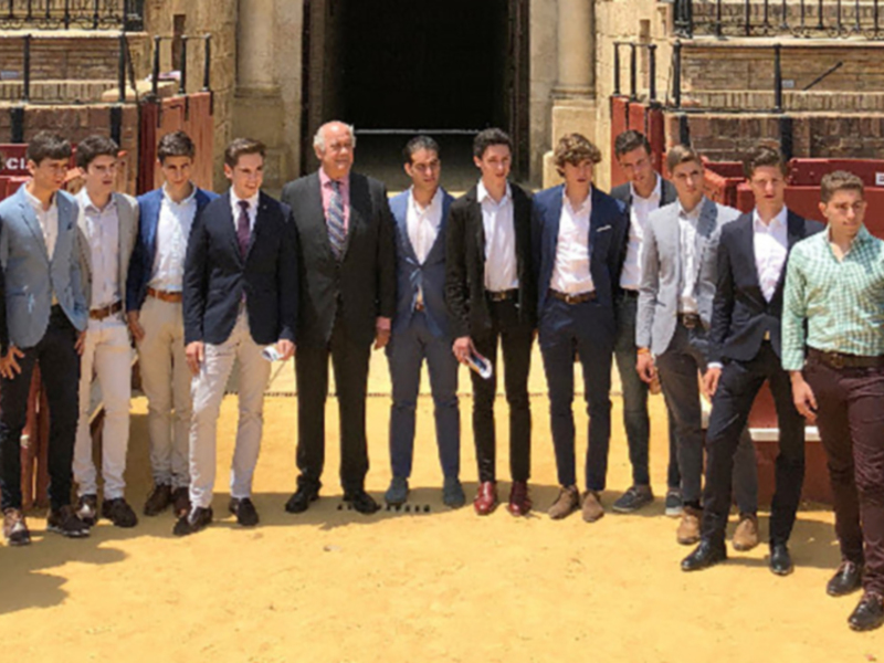 Un grupo de jóvenes hombres vestidos con trajes y camisas blancas posan en una plaza de toros, con una estructura de madera y un fondo de arena roja.