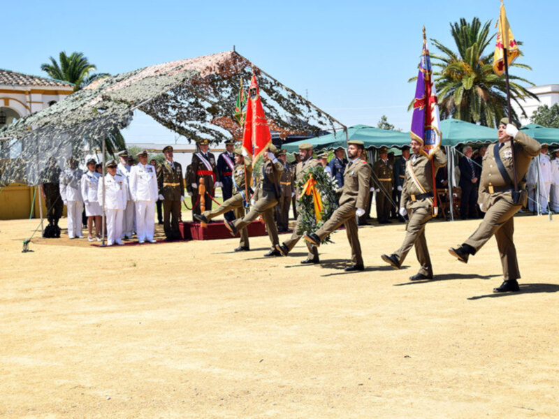 Desfile militar con soldados marchando bajo un toldo de camuflaje y banderas en un escenario al aire libre.