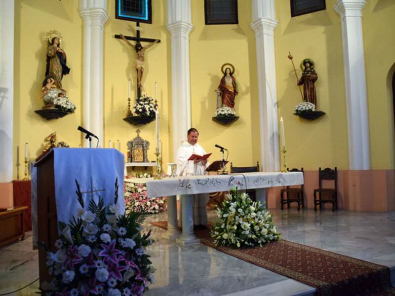 Interior de una iglesia con altar adornado con flores, estatuas religiosas y un sacerdote leyendo un libro. La decoración es elegante y simbólica, reflejando la importancia religiosa de la escena.
