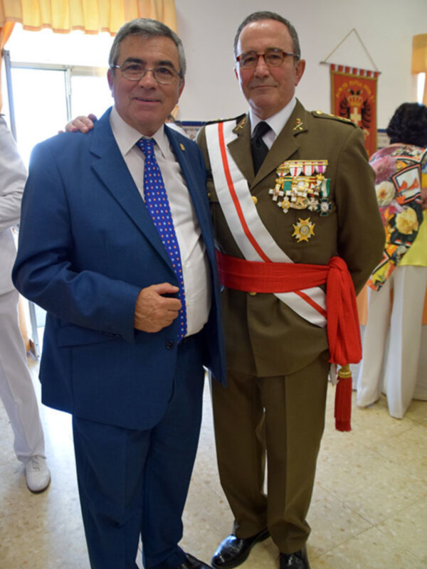 Dos hombres en trajes formales, uno en uniforme militar con medallas y sash rojo, posan juntos en una sala decorada.
