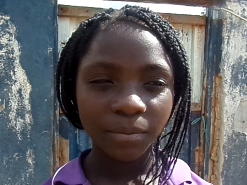 Una niña con trenzas de color negro, mirando directamente a la cámara, con un fondo de paredes azul y blanca en un entorno urbano.