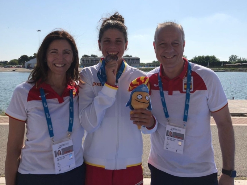 Tres atletas celebran juntos con medallas y una mascota, posando al aire libre junto a un lago.