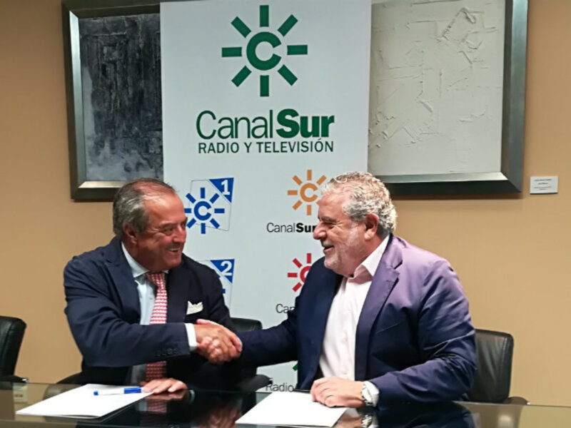 Dos hombres en trajes se saludan con una sonrisa, sentados en una mesa con un fondo que incluye el logo de Canal Sur Radio y Televisión.