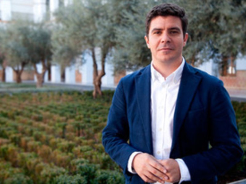 Hombre en traje azul y camisa blanca, posando al aire libre con árboles y arbustos en el fondo.