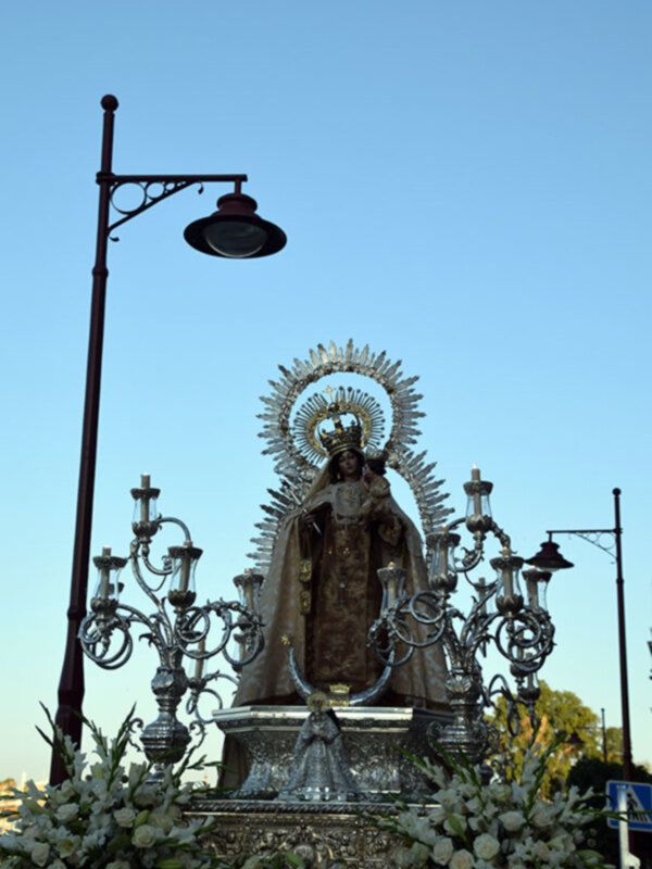 Procesión religiosa con estandarte de Virgen María adornado con flores y candelabros, bajo un cielo claro.