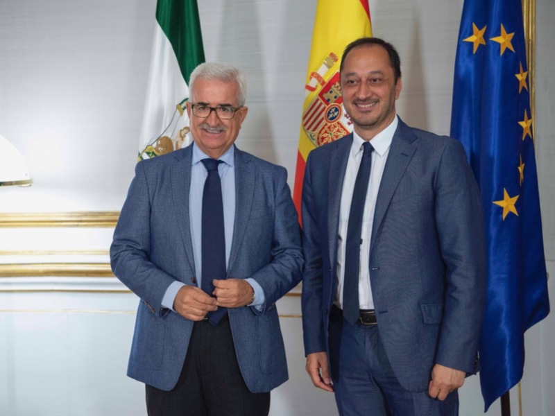 Dos hombres en trajes oficiales posan frente a banderas de España, la Unión Europea y México.