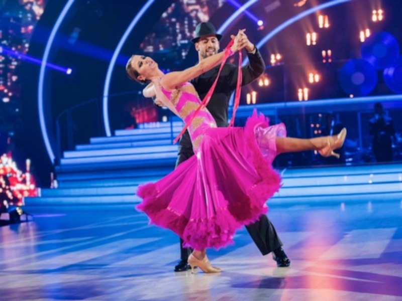 Baile latino con pareja en escenario iluminado, vestida con vestido rosa y el hombre en traje negro y sombrero.