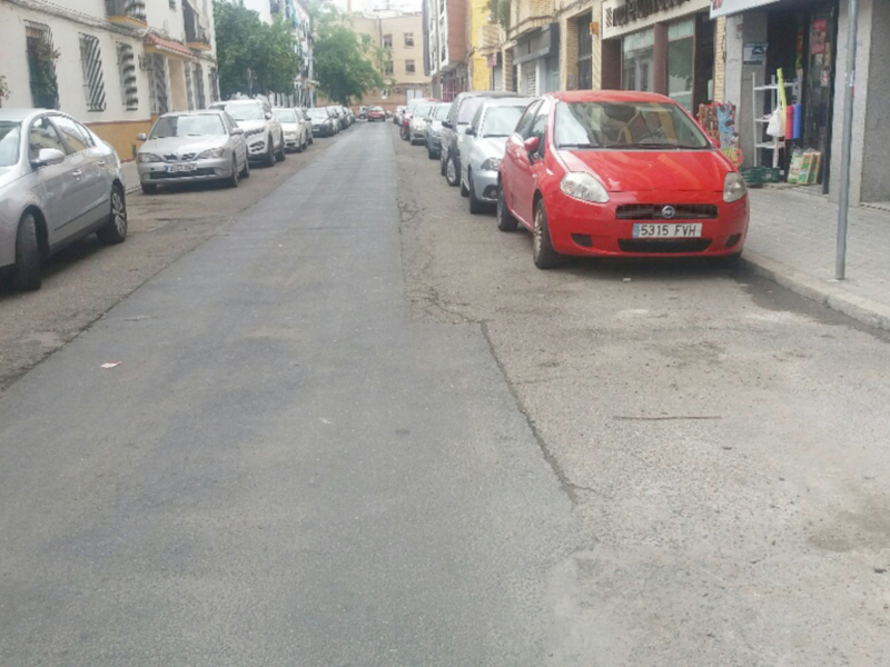 Vehículos aparcados en una calle urbana con tiendas y edificios en el fondo.