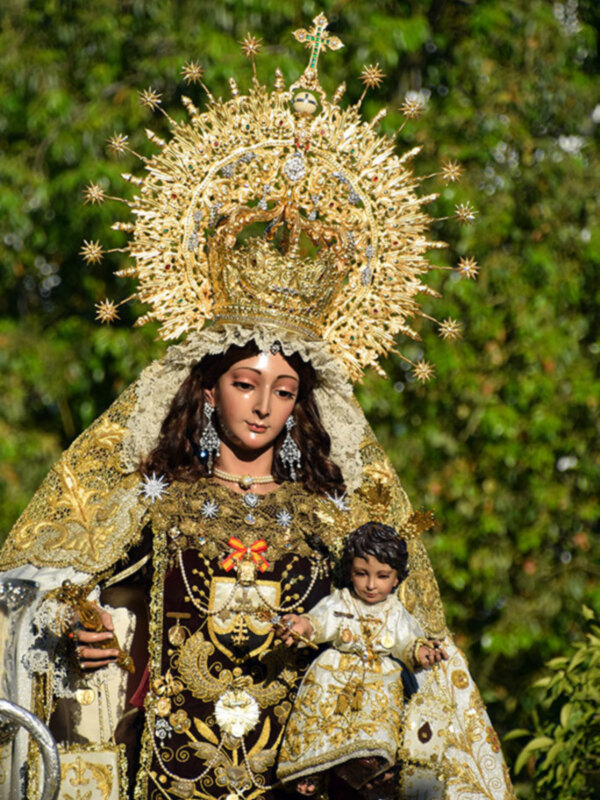 Estatua de la Virgen con un niño en sus brazos, vestida con una capa dorada y joyas. Fondo de árboles verdes.