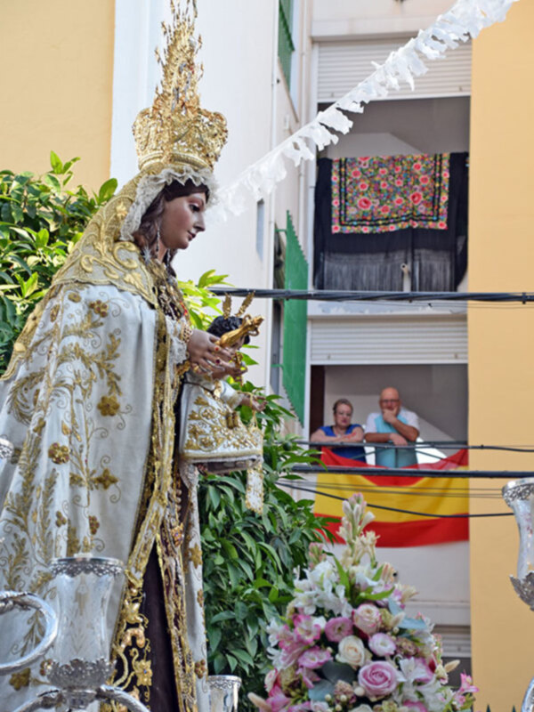 Una imagen de una figura vestida con trajes tradicionales, posiblemente una estatua religiosa o personaje histórico, con un fondo que muestra una ventana con cortinas y una bandera española.