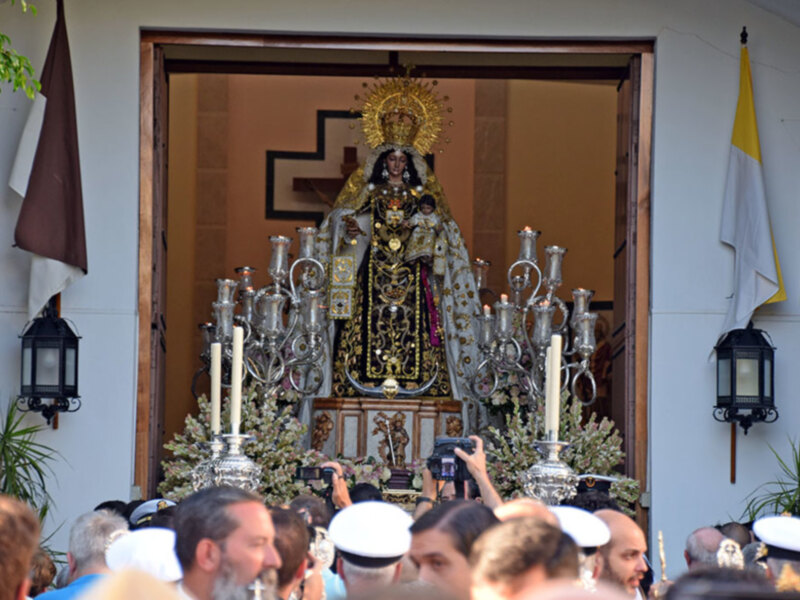 Imagen de una procesión religiosa con una imagen venerada en el centro, rodeada por gente y velas encendidas. Banderas y decoraciones religiosas en el fondo.