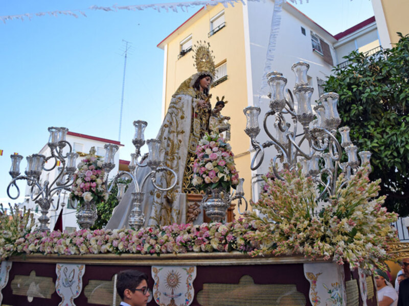 Imagen de una procesión religiosa con una imagen de la Virgen en un carro adornado con flores y candelabros. Fondo de edificios y cielo azul claro.