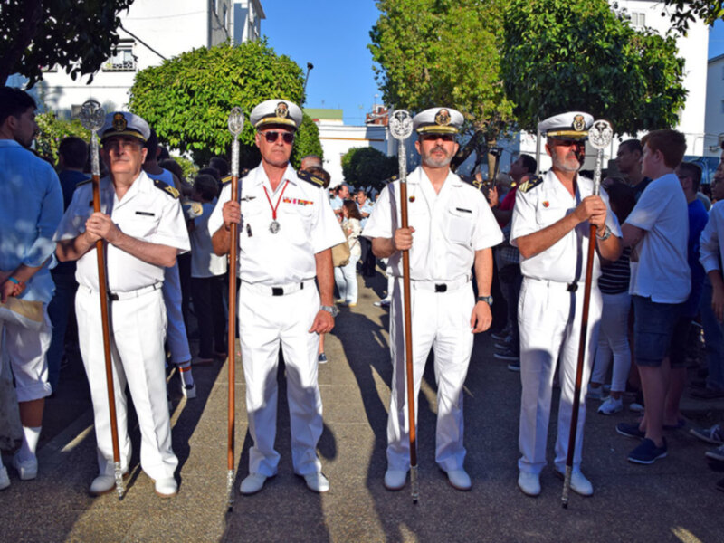 Tres hombres vestidos con uniformes militares blancos y sombreros de ala azul, posan en una calle con árboles verdes y edificios blancos al fondo. El suelo es de asfalto y hay otros personas en el fondo, algunas con sombrillas.