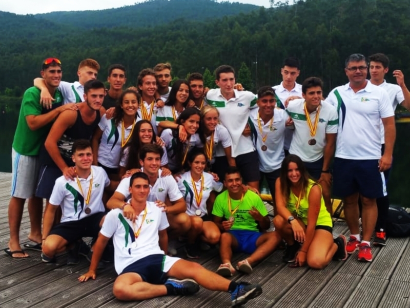 Equipo deportivo con medallas posando juntos en un muelle junto a un lago, con un paisaje de montañas en el fondo.