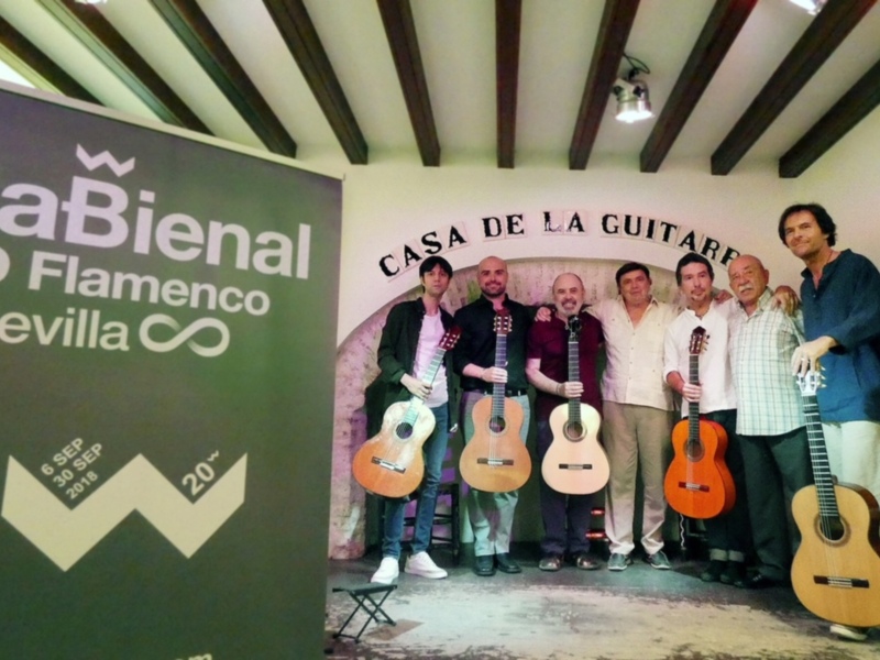 Grupo de músicos posando con sus guitarras en una sala con decoración de madera y un cartel que anuncia una bienal de flamenco en Sevilla en 2018.