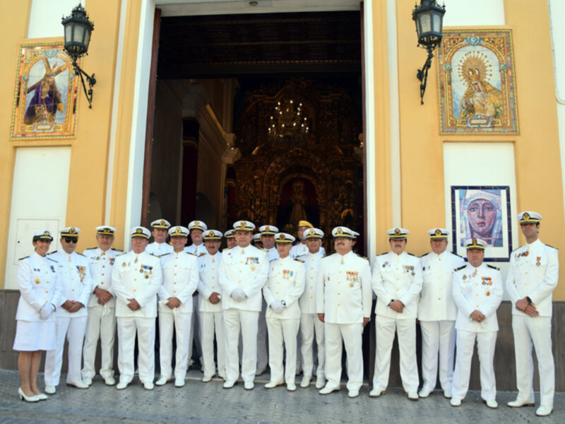 Fotografía de oficiales militares en uniforme blanco frente a una iglesia con una imagen de la Virgen María en el muro.