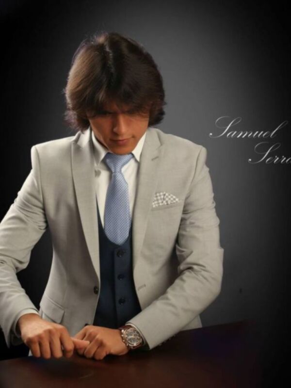 Elegante hombre en traje formal con corbata y reloj, sentado en un escritorio oscuro.
