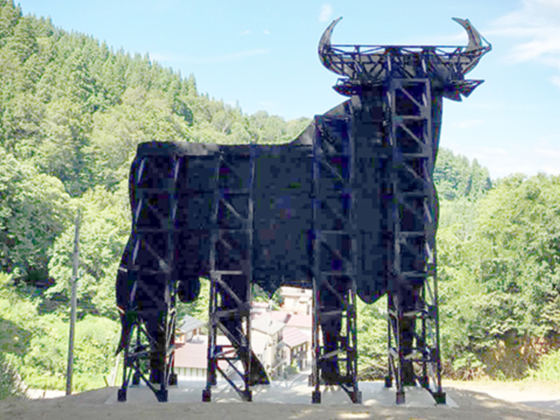 Escultura monumental de un toro en un entorno natural, rodeada de árboles y con un cielo azul claro.