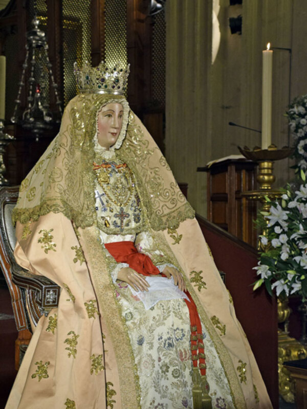 Imagen de una imagen de la Virgen María vestida con un manto y corona, sentada en un trono en un interior de iglesia con elementos religiosos alrededor.