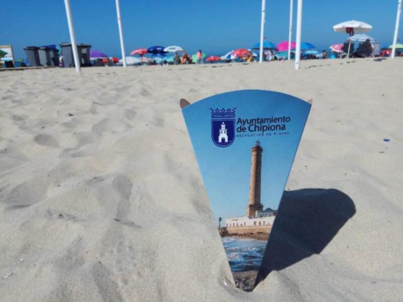 Playa con sombrillas y palmeras, con un cielo azul claro y una torre en la distancia.
