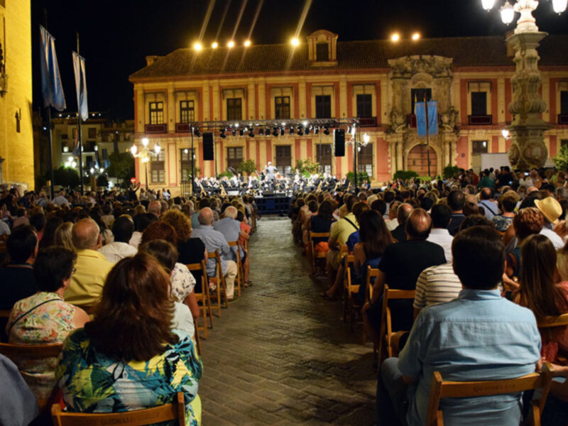 Concierto al aire libre en un edificio histórico con público sentado en sillas al aire libre bajo luces de teatro.