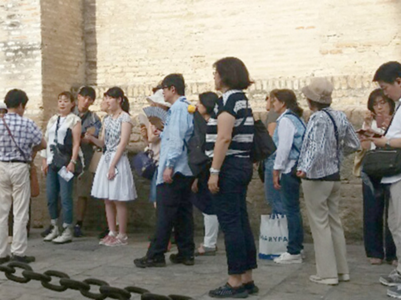 Un grupo de personas camina junto a una pared de piedra, con algunas llevando bolsas y otros intercambiando conversaciones.