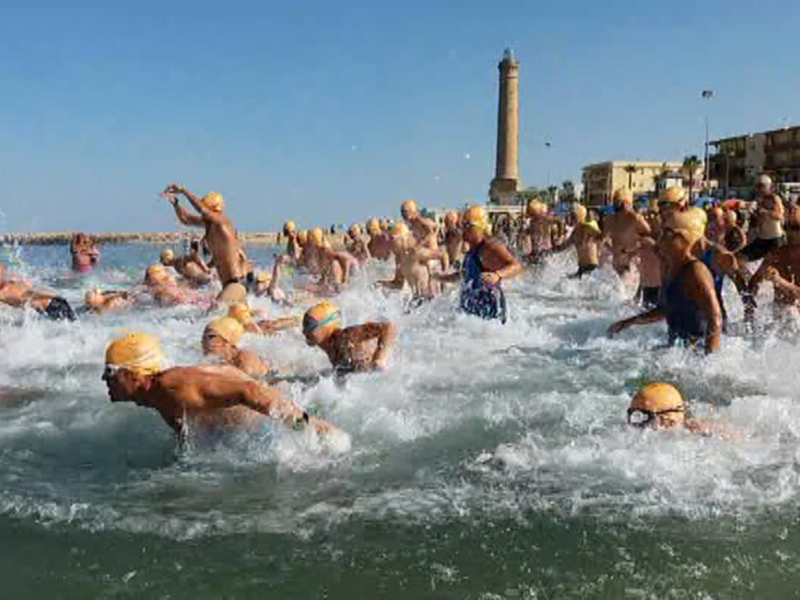Una multitud de nadadores en la playa, con un faro en el fondo bajo un cielo azul claro.