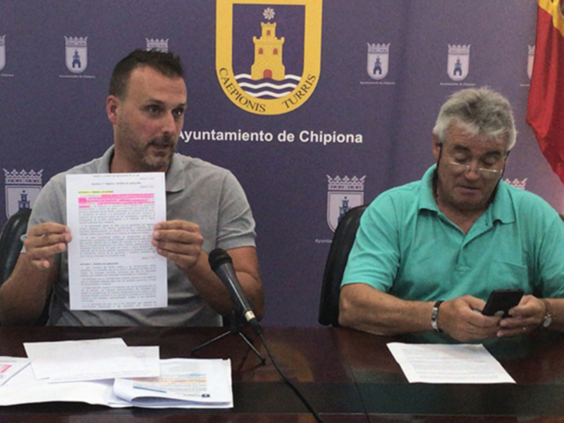 Conferencia de prensa en Chipiona, con representantes del Ayuntamiento y bandera española en el fondo.