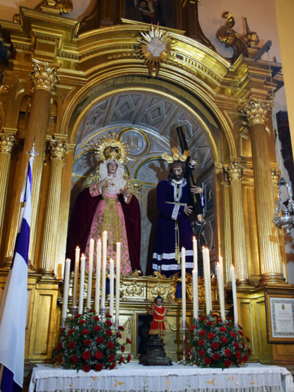 Altar de una iglesia con estatuas religiosas y velas encendidas, decorado con flores y un manto dorado.