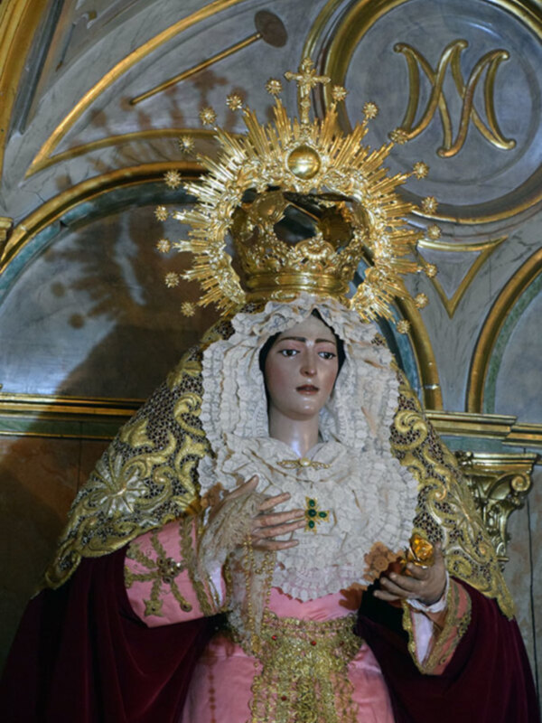 Estatua de la Virgen María con vestimentas y corona dorada en un entorno religioso.